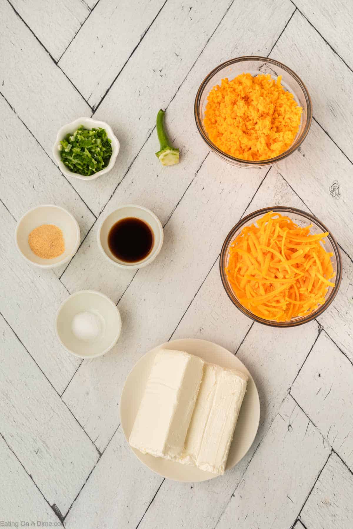 Top view of various ingredients on a white wooden surface: two blocks of cream cheese on a plate, shredded cheese, diced jalapeños, shredded cheddar cheese, a small bowl of soy sauce, garlic powder, and a small bowl of salt — perfect for crafting a Pumpkin Cheeseball.