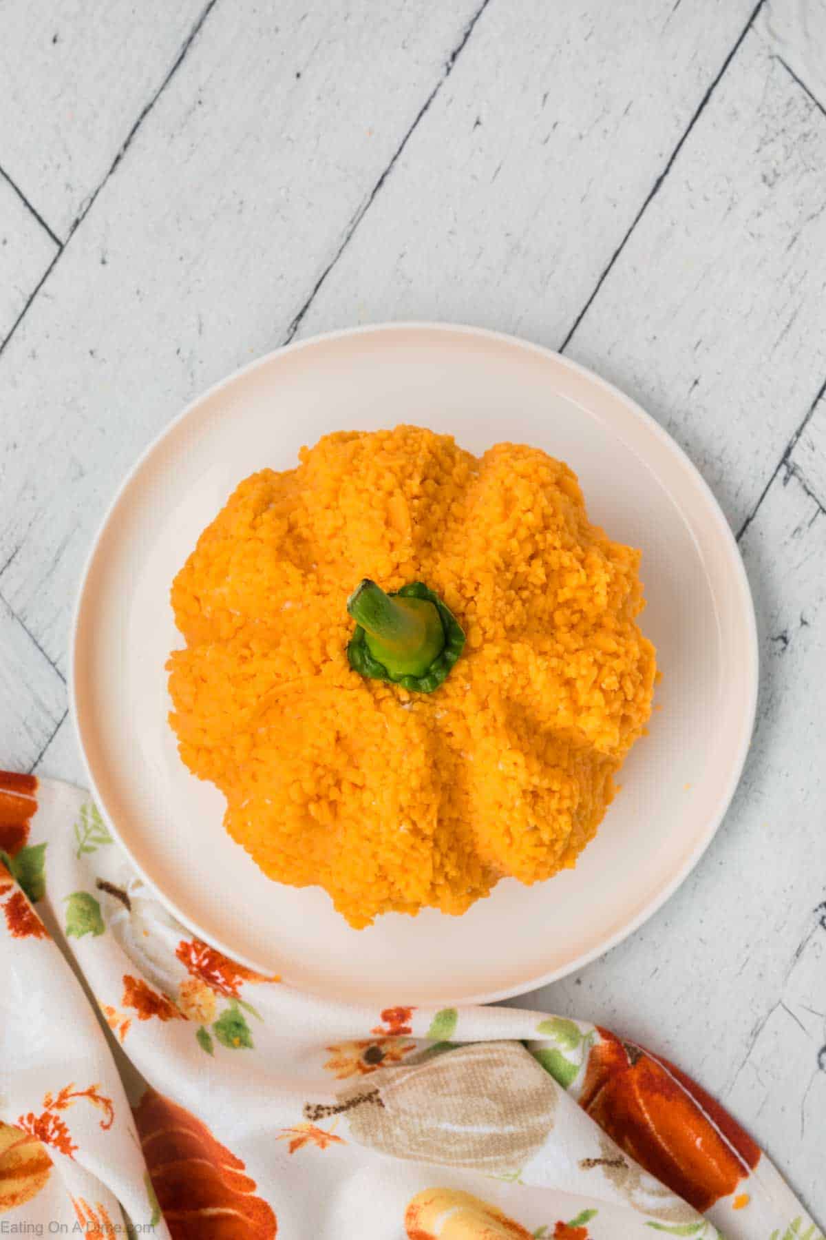 A round plate sits on a rustic white wooden surface, displaying a serving of rice shaped like a pumpkin. The orange rice mound mimics the ridges of a pumpkin, with a small green garnish on top to resemble the stem. Nearby, a patterned cloth is partially visible next to what looks like an appetizing cheeseball.