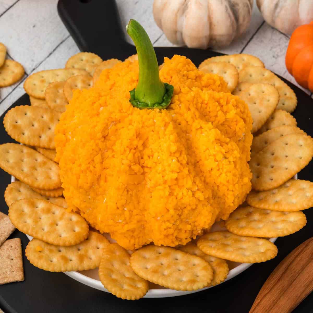 A Pumpkin Cheeseball shaped like a pumpkin, covered in finely crushed cheese bits with a green stem on top, is placed on a white plate surrounded by crackers. The plate is on a black serving board, with pumpkins in the background.