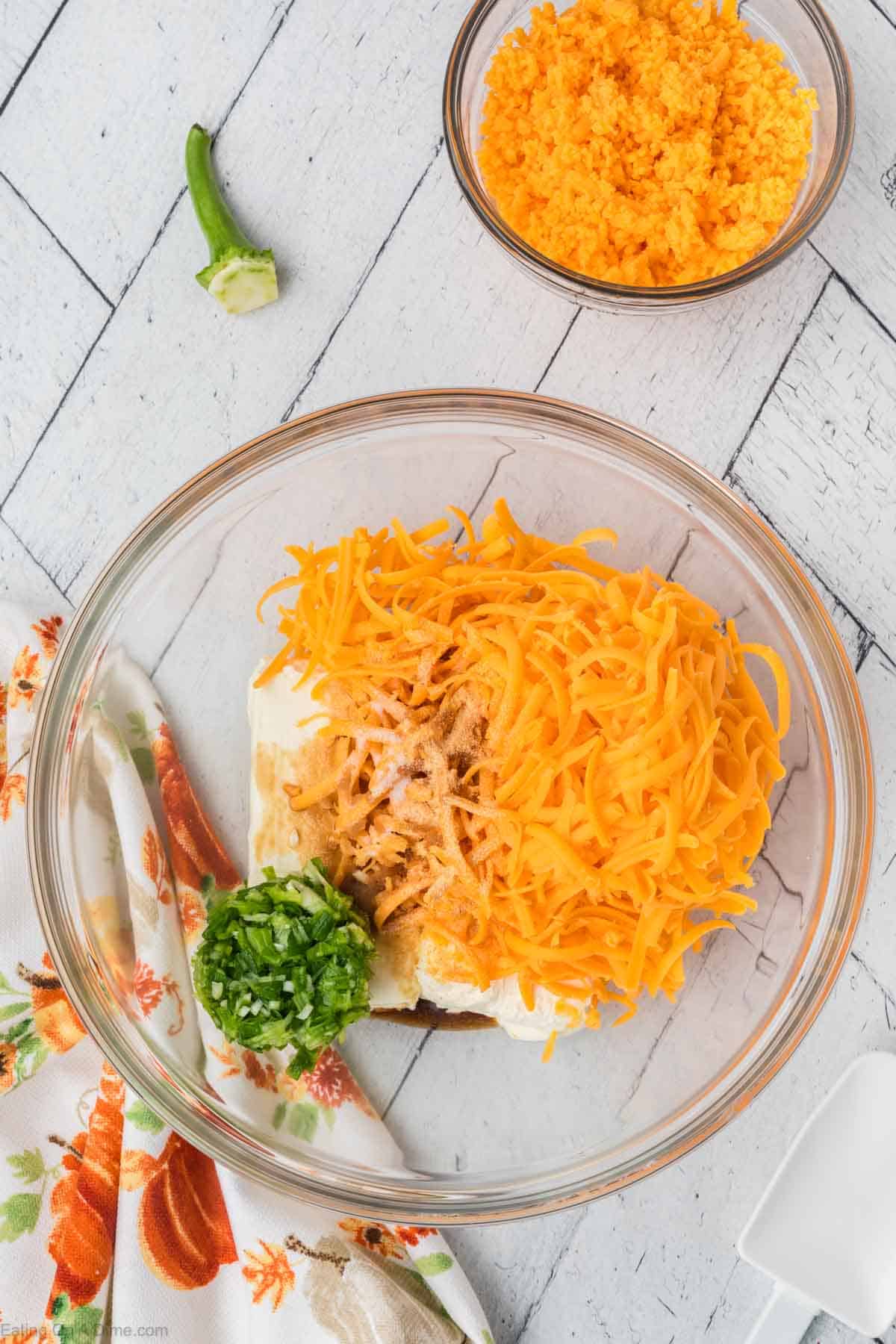 A clear glass bowl on a white wooden surface contains shredded cheddar cheese, cream cheese, chopped green peppers, and spices—perfect for crafting a Pumpkin Cheeseball. Next to the bowl is a floral-patterned napkin. In the background, another bowl with shredded cheddar cheese and a sliced jalapeño are visible.