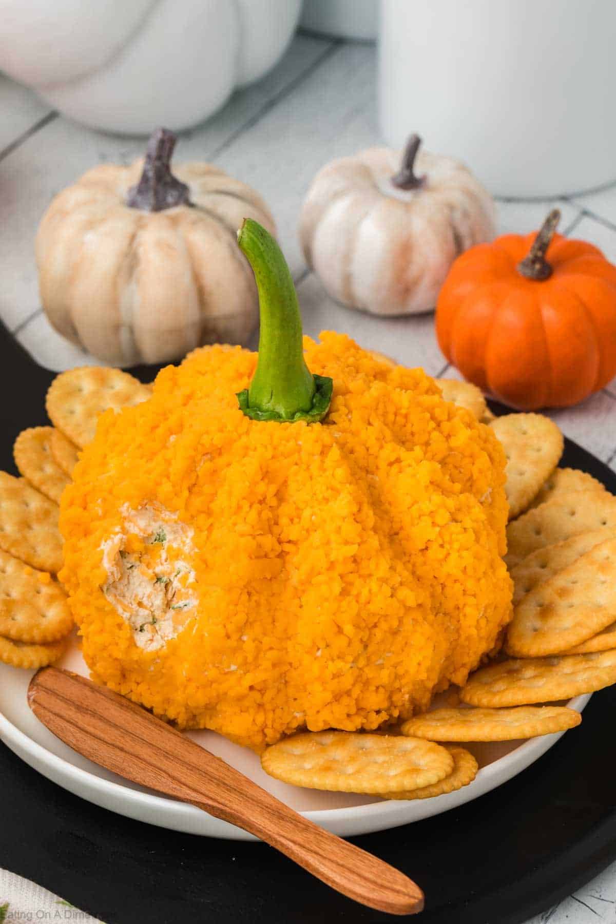 An orange, pumpkin-shaped cheeseball with a green stem is placed on a white plate surrounded by round crackers. The cheeseball has a small portion scooped out, revealing a creamy filling. In the background, there are white and small orange pumpkins.