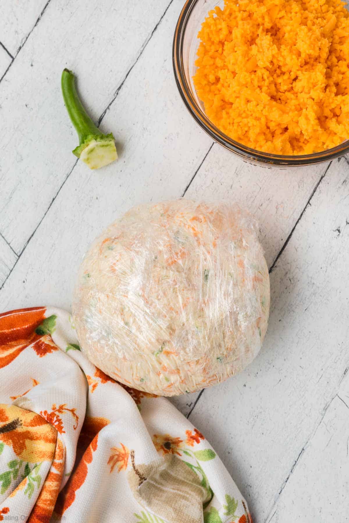 A cheeseball of dough wrapped in plastic wrap lies on a white wooden surface next to a bowl of grated orange cheese and a green chili pepper with its top cut off. A colorful cloth with a floral print is partially visible in the corner.