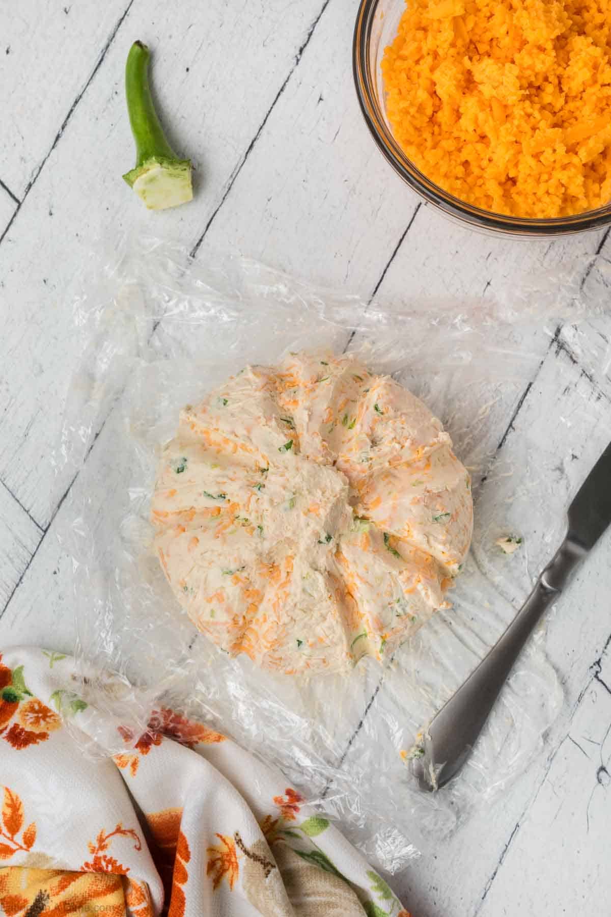 A delectable pumpkin cheeseball with herbs sits on clear plastic wrap on a white wooden surface. A partially cut green pepper lies nearby, while a knife rests beside the cheeseball. In the upper right, a bowl of grated cheese can be seen. A floral patterned cloth is partially visible.