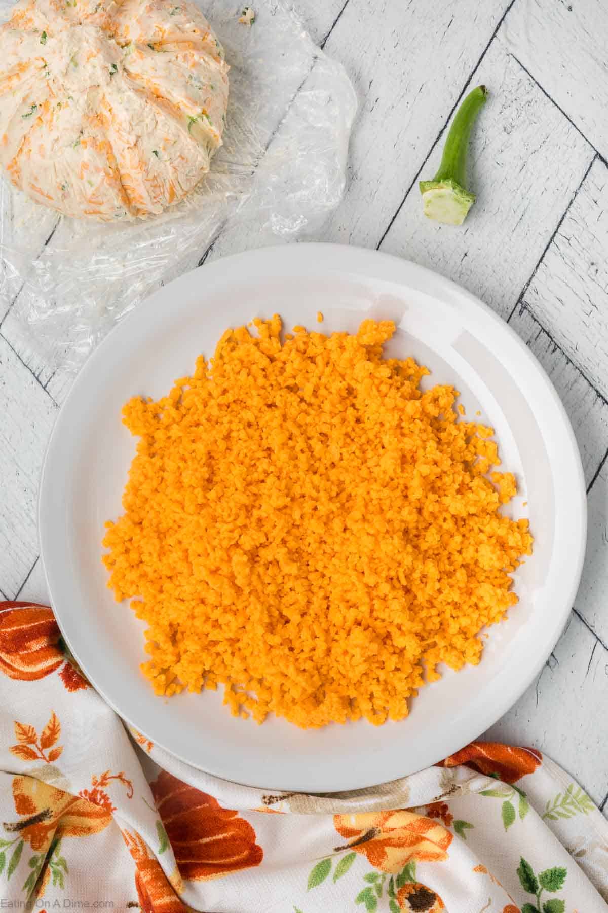 A white plate filled with vibrant orange cauliflower rice, resembling the hues of a pumpkin, placed on a light wooden table. Beside the plate is a partially unwrapped plastic package of cream cheese and a fresh green asparagus spear. A white and orange patterned cloth is draped nearby.