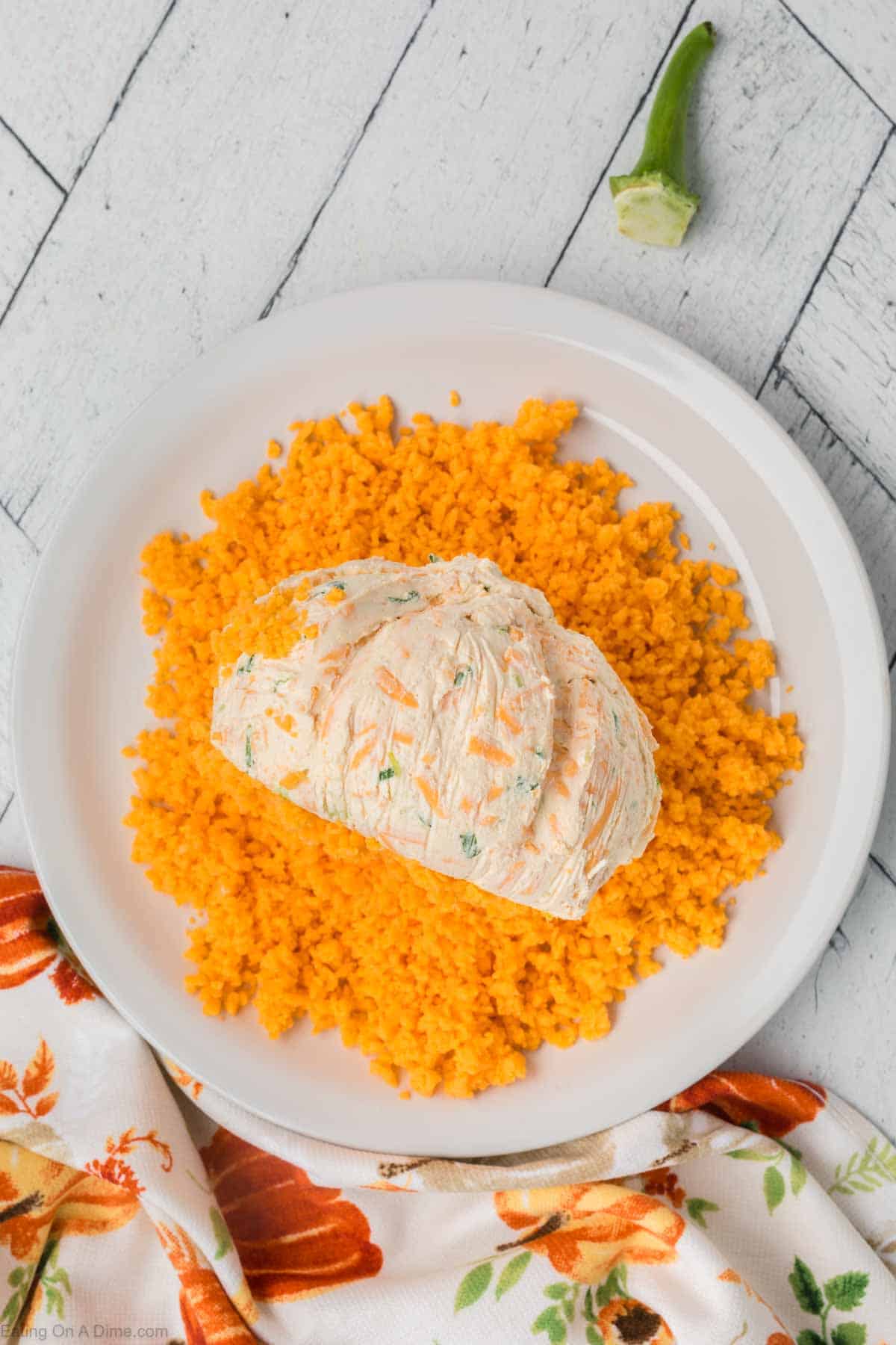 A white plate holds a generous serving of yellow-colored rice, topped with a scoop of herb and vegetable cream cheese, resembling a pumpkin cheeseball. The plate rests on a wooden surface, accompanied by a patterned cloth and a small green pepper stem nearby.