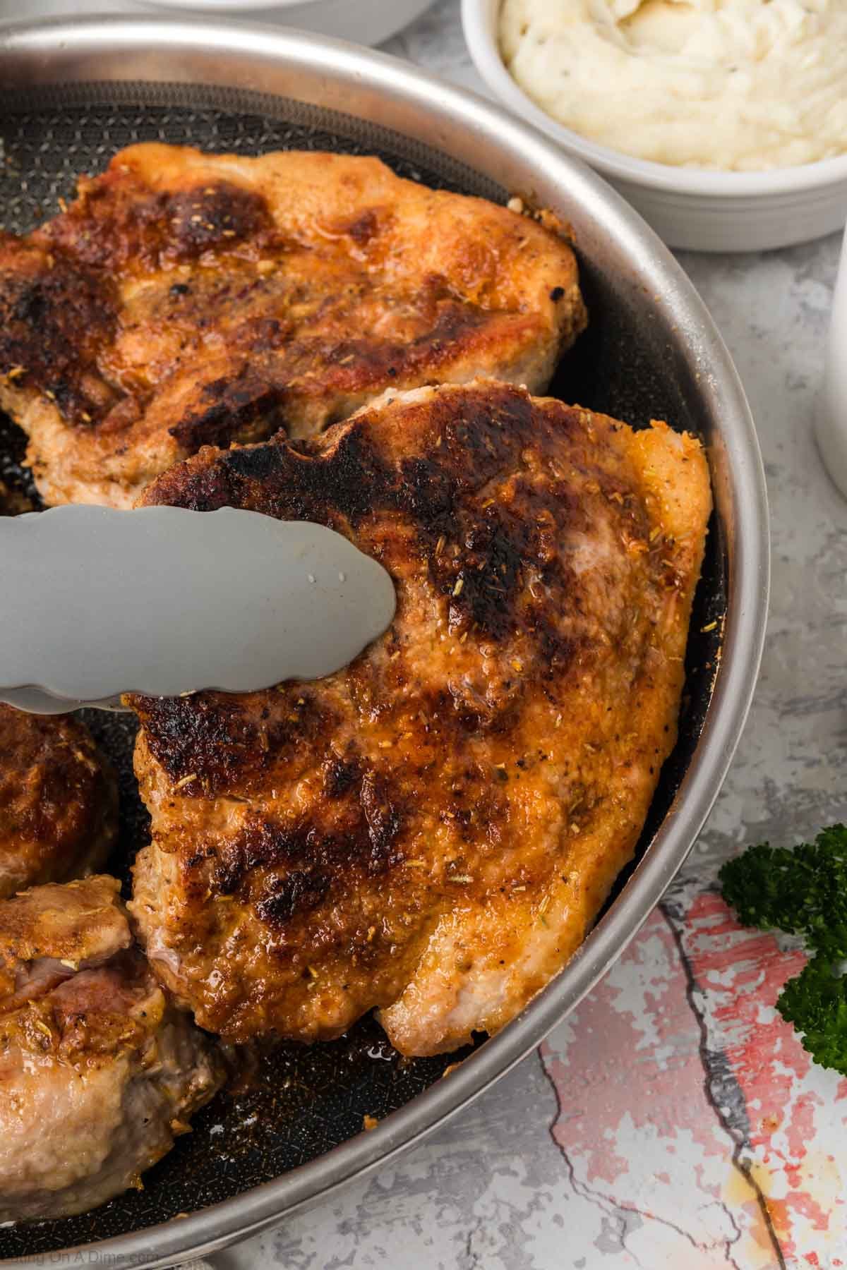 Golden brown pork chops in a skillet with a pair of tongs flipping one day