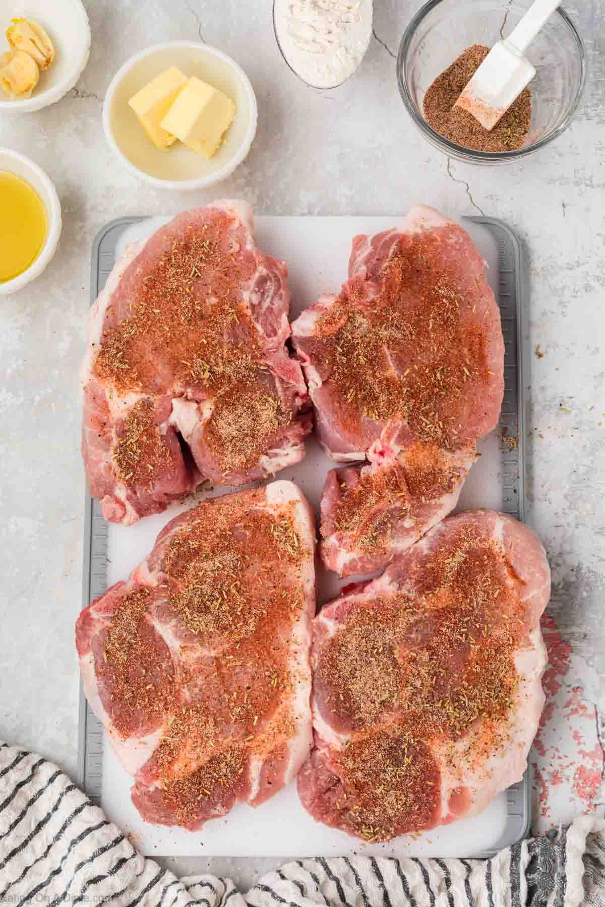 Seasoning spread over raw pork chops on a cutting board with small bowl of butter and seasoning