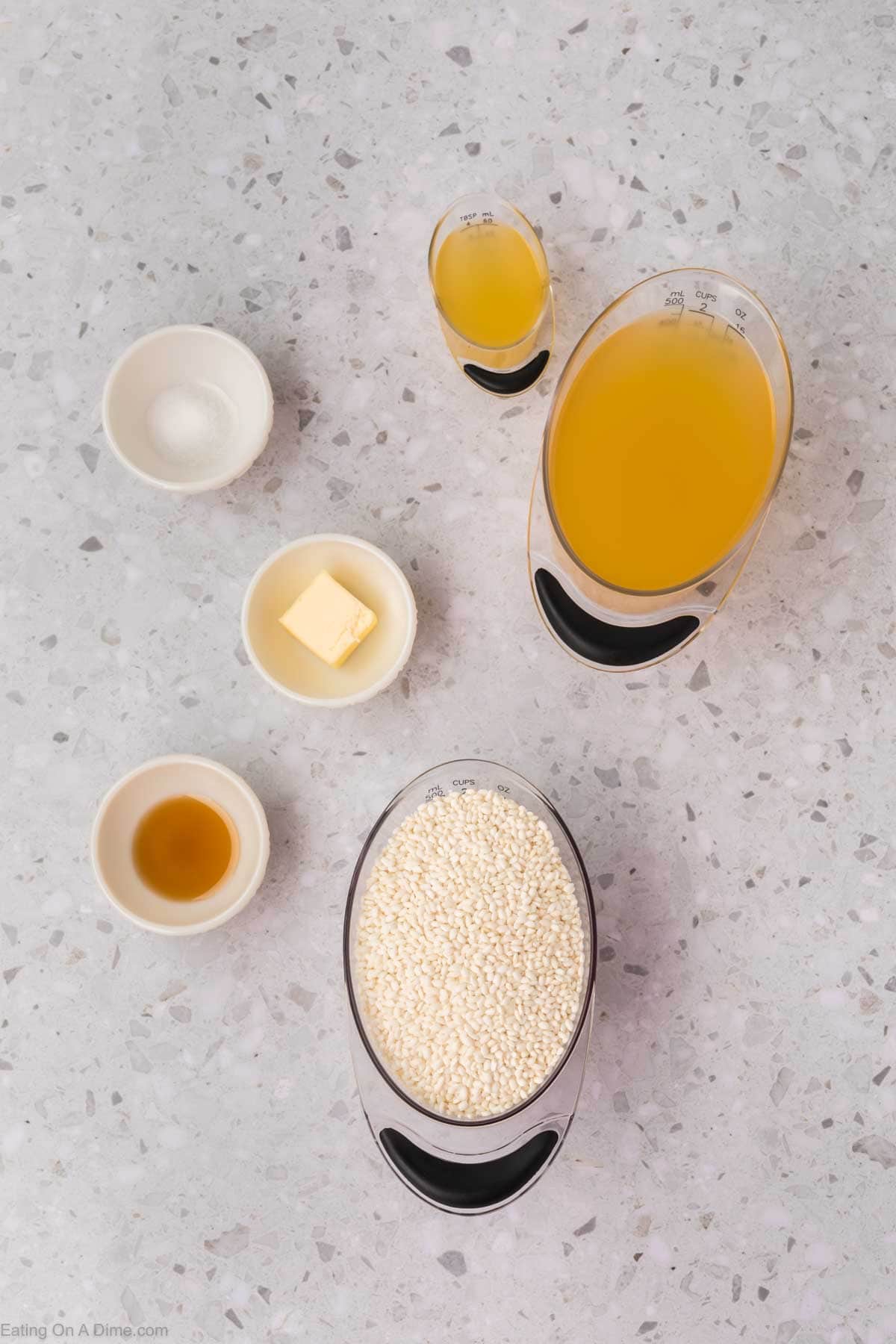 A top-view image displays ingredients in various bowls and measuring cups on a speckled countertop. Visible are chicken broth, butter, Arborio rice, sticky rice, white wine, salt, and olive oil. The items are neatly arranged and ready for cooking.