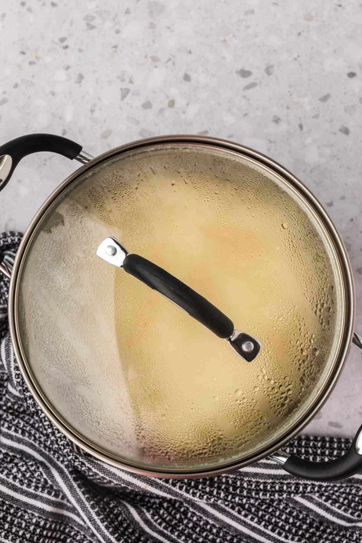 A pot with a glass lid containing boiling sticky rice is placed on a gray countertop. Steam is visible on the lid. The pot has black handles, and a striped black and white kitchen towel is partially wrapped around its base.