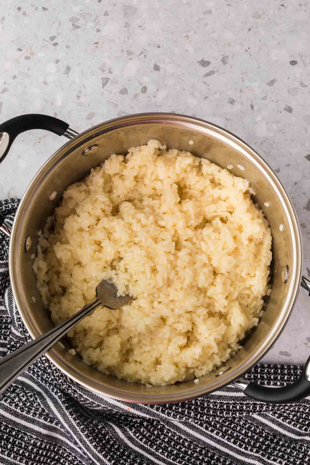 A stainless steel pot filled with sticky rice is placed on a gray speckled countertop. A silver fork is sticking out of the rice. The pot rests on a black and white striped cloth.