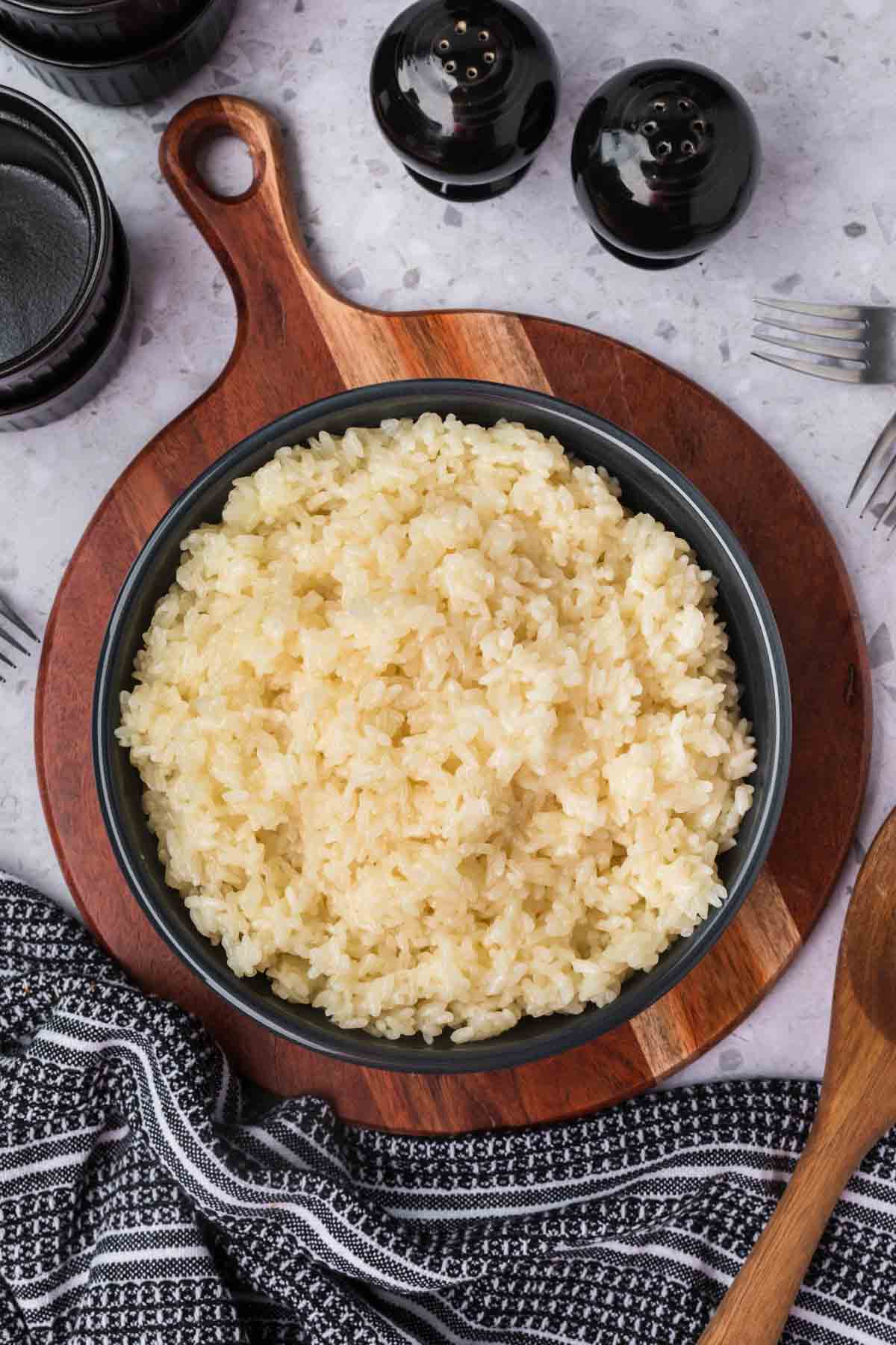 A bowl of sticky rice placed on a round wooden cutting board. The setup includes black pepper and salt shakers, a few forks, a wooden spoon, and a striped cloth napkin on a speckled countertop.