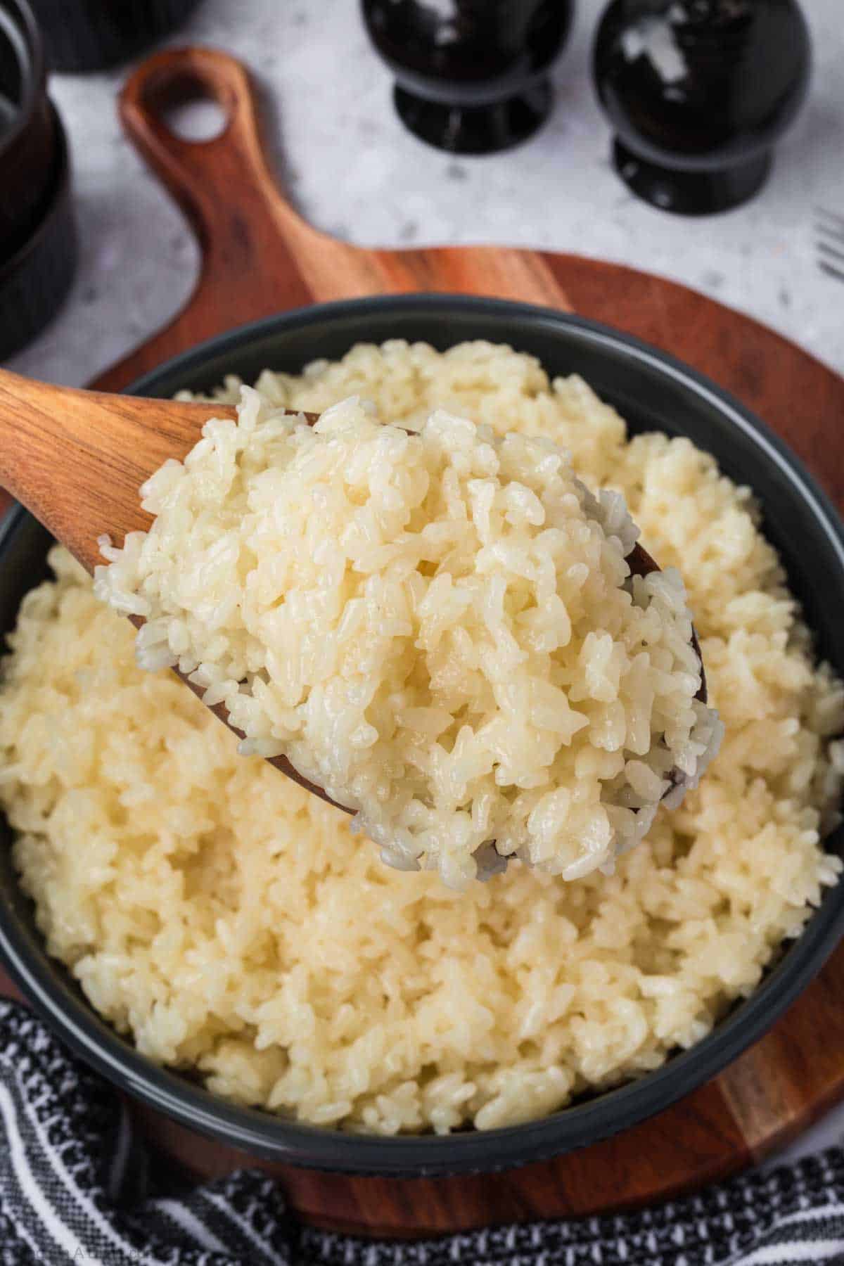 A wooden spoon holds a serving of fluffy, sticky rice above a bowl filled with more rice. The bowl rests on a wooden cutting board, and in the blurred background, black salt and pepper shakers stand. A striped cloth is partially visible in the corner.