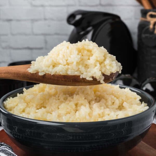 A wooden spoon holding a portion of sticky rice is positioned above a dark bowl filled with more rice. The background includes a white brick wall and assorted kitchen items, slightly out of focus.