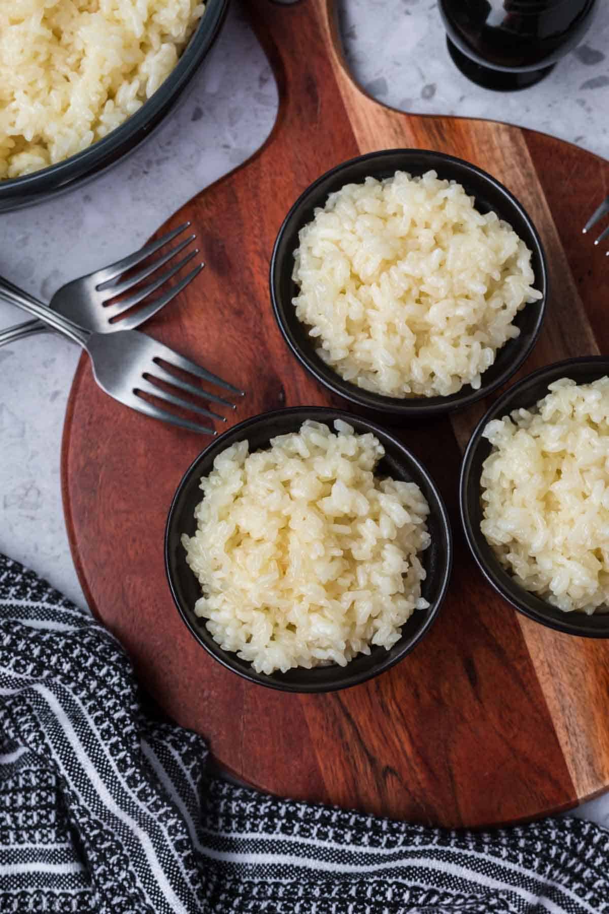 Top view of three small bowls filled with cooked sticky rice placed on a wooden board. Two forks are nearby, along with a textured black and white cloth. Part of a larger bowl with more rice is visible in the corner.