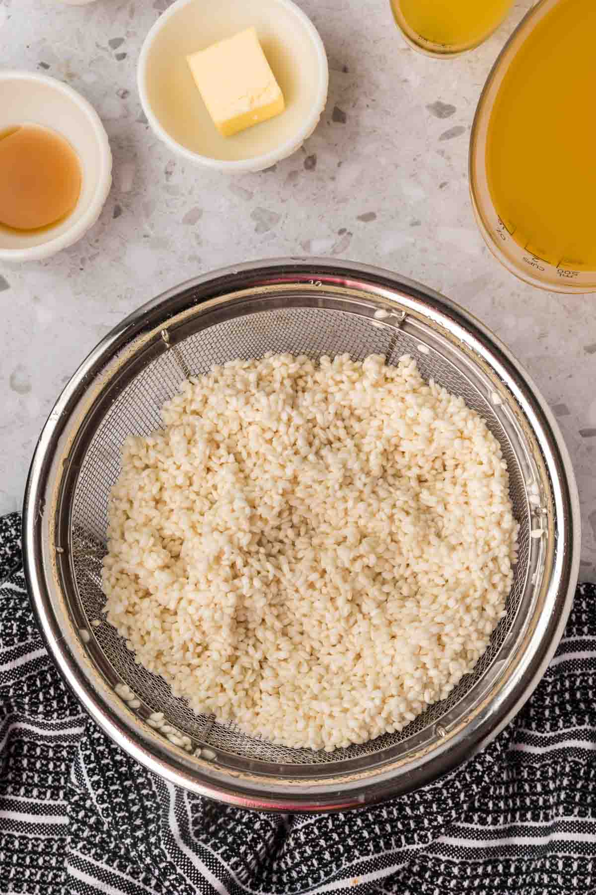 A steel colander filled with rinsed, uncooked sticky rice sits on a marble countertop. Surrounding it are small bowls containing butter, broth, and another liquid ingredient. A dark patterned cloth is partially visible at the bottom of the image.