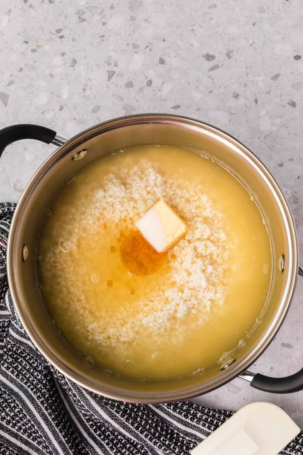 A pot on a grey-speckled countertop contains bubbling yellow liquid with a small pad of butter melting on top of sticky rice. The pot has a black handle, and there is a black and white striped cloth partially visible beneath it.