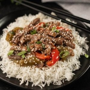 A black plate with a serving of white rice topped with stir-fried beef, red bell peppers, green peppers, and chopped green onions, all garnished with sesame seeds. Black chopsticks and a black-and-white checkered napkin are in the background—a perfect dish for your Meal Plan 24.