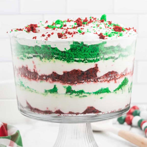 A Christmas trifle in a glass dish features layers of red and green cake, fluffy white frosting, and crumbled cake pieces on top, set against a white tiled background. A striped napkin and some marshmallows are partially visible nearby, adding to the festive charm.