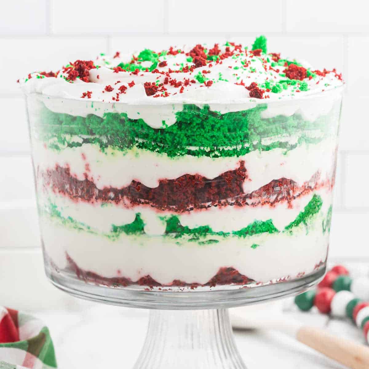 A Christmas trifle in a glass dish features layers of red and green cake, fluffy white frosting, and crumbled cake pieces on top, set against a white tiled background. A striped napkin and some marshmallows are partially visible nearby, adding to the festive charm.