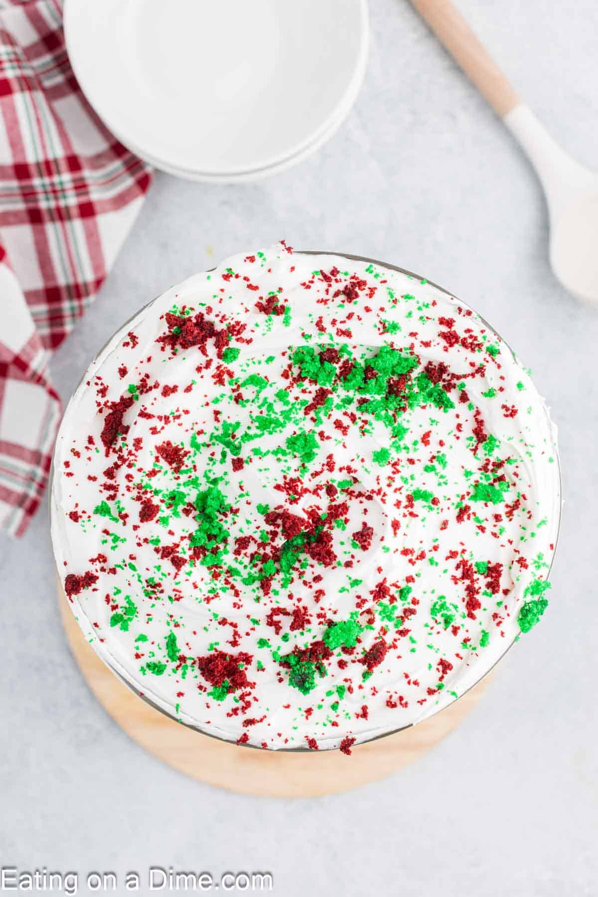 A festive trifle dessert sits in a bowl, topped with white frosting and sprinkled with red and green cake crumbs, capturing the essence of Christmas. Nearby, a plaid cloth adds a cozy touch, accompanied by a white spoon and neatly stacked empty bowls.