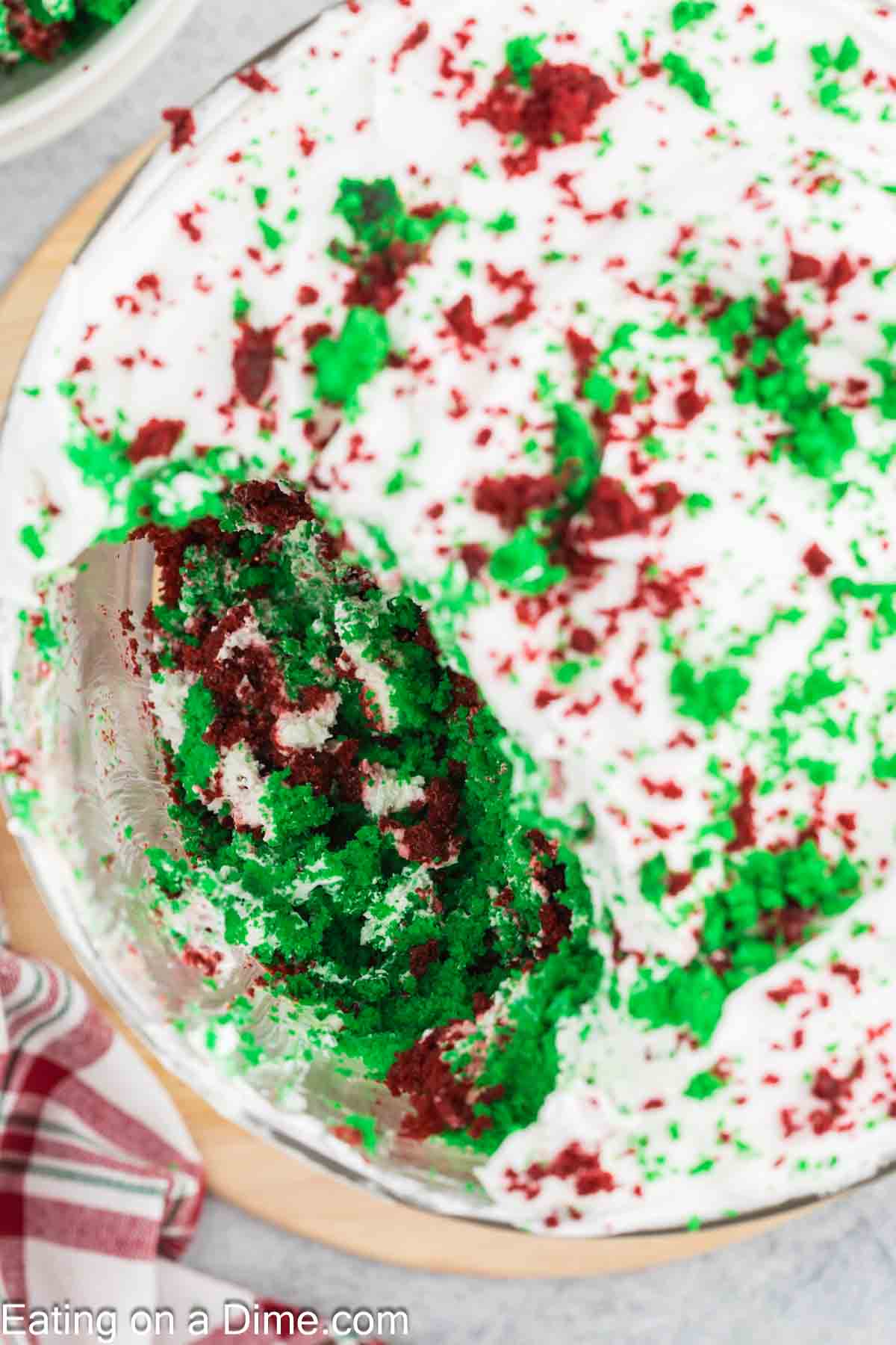 A festive Christmas cake adorned with red and green sprinkles on white frosting, partially sliced to reveal trifle-like layers of red, green, and white inside. It's elegantly placed on a table with a red and white striped cloth nearby.