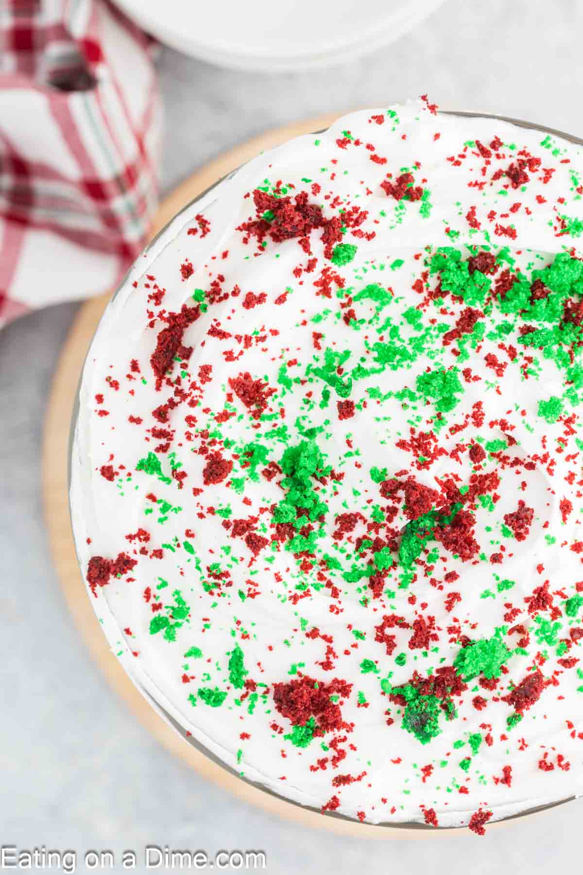 A close-up of a festive Christmas cake topped with white frosting and sprinkled with green and red crumbles, reminiscent of a holiday trifle. A red and white plaid napkin is visible in the background.