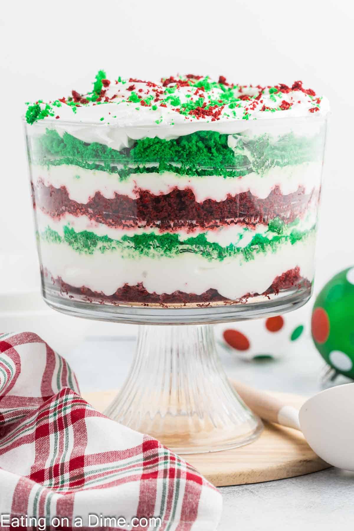 A festive Christmas trifle in a glass dish with red velvet cake, green cake, and creamy white layers. Topped with whipped cream and red and green sprinkles. A red and green plaid cloth and decorative balls set the merry background perfectly.