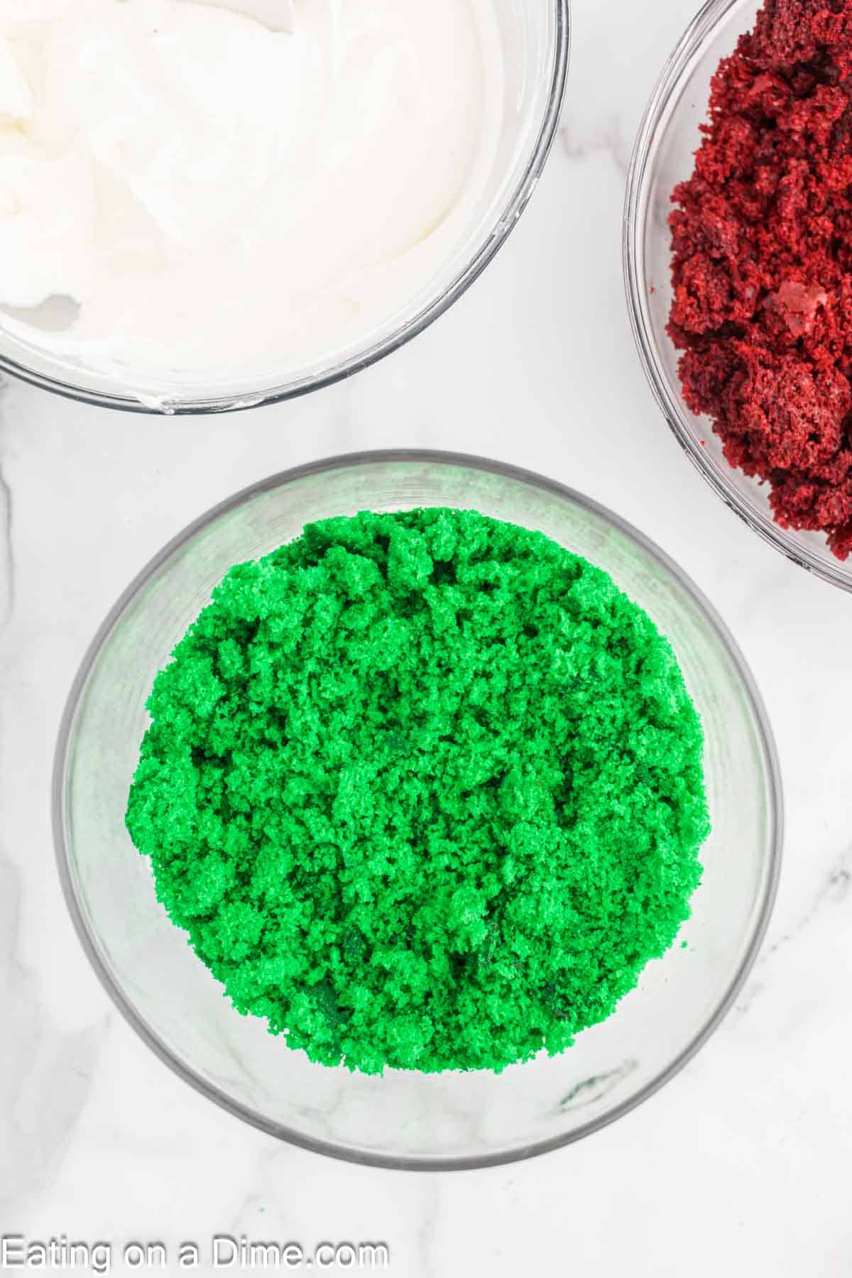 Three bowls sit elegantly on the marble surface: one with rich white frosting, another brimming with vibrant red cake crumbs, and a third showcasing lively green cake crumbs, reminiscent of a festive Christmas trifle.