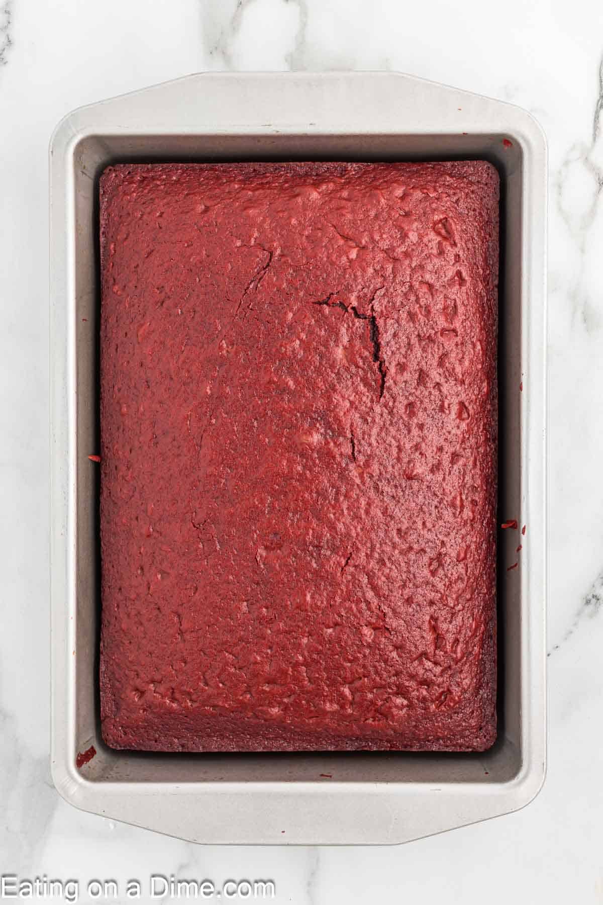 A freshly baked red velvet cake rests in a rectangular metal pan on a marble surface, evoking festive Christmas cheer. The cake's surface has a slight crack in the center, and it appears moist and even in texture.