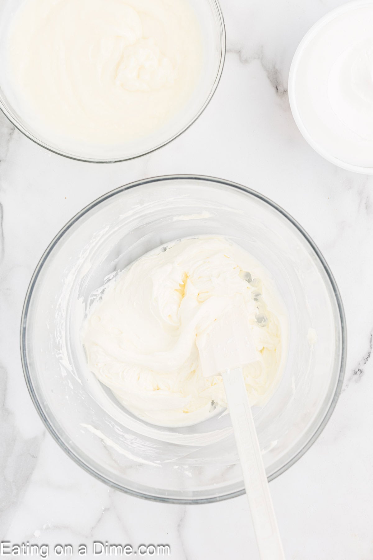 A clear glass bowl filled with a creamy, white cheese mixture resembles the makings of a delightful Christmas trifle, with a white spatula resting in it. Nearby, two other bowls hold similar decadent mixtures on a pristine white marble surface.