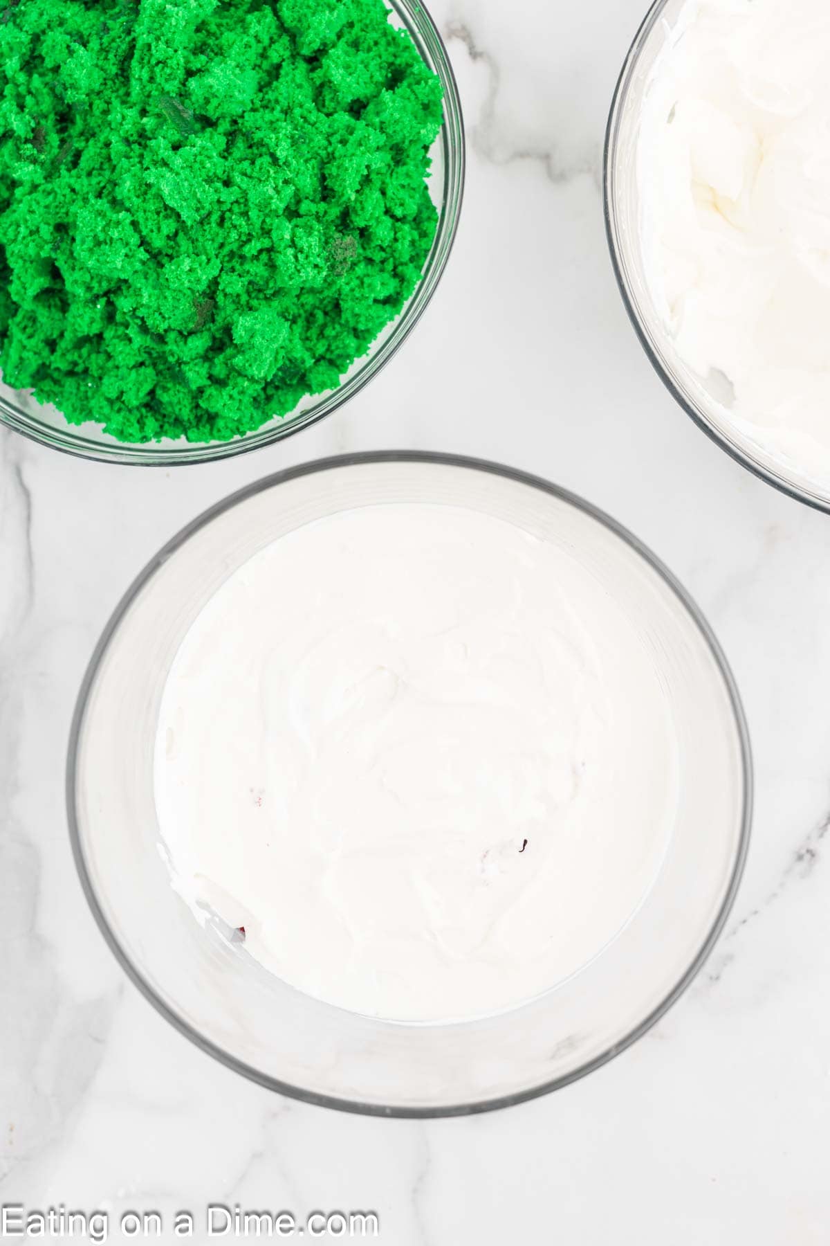 Three bowls on a marble surface: one with green cake crumbs, another with white frosting, and a third with more frosting, ready to create a festive Christmas trifle.