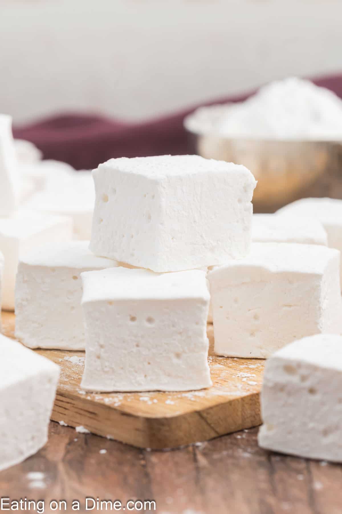 Close-up of marshmallows stacked on a wooden board. These homemade treats are white, soft, and have a fluffy texture. A bowl with powdered ingredients sits in the background, highlighting the artisanal touch.