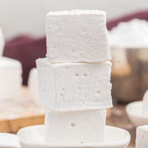 Three square homemade marshmallows stacked on a small plate. The background features a blurred bowl and additional marshmallows, creating a warm, inviting atmosphere perfect for enjoying your favorite marshmallow recipe.