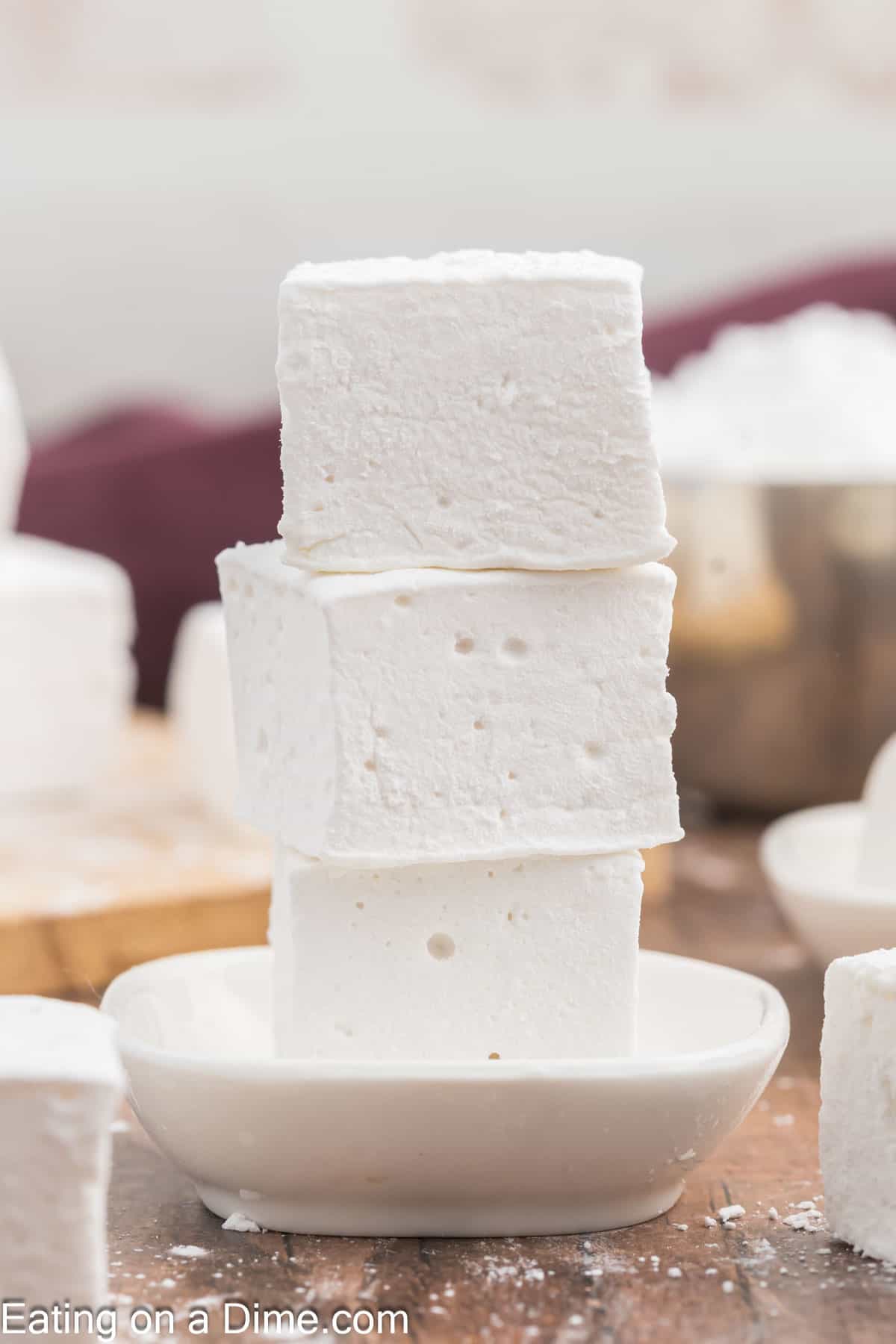 A stack of three large, fluffy white homemade marshmallows is balanced on a small dish. The cozy background, with more marshmallows and a bowl, hints at DIY marshmallow fun and sweet creations in the making.