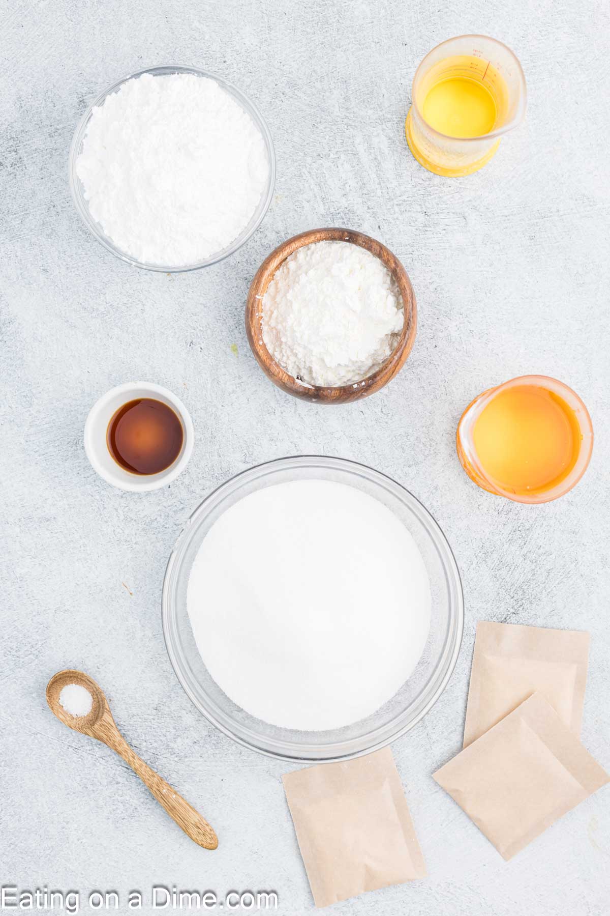 Various baking ingredients are arranged on a light gray surface, perfect for homemade marshmallows, including powdered sugar, corn syrup in jars, vanilla extract in a small bowl, and baking powder in paper packets. A wooden spoon rests beside the items.