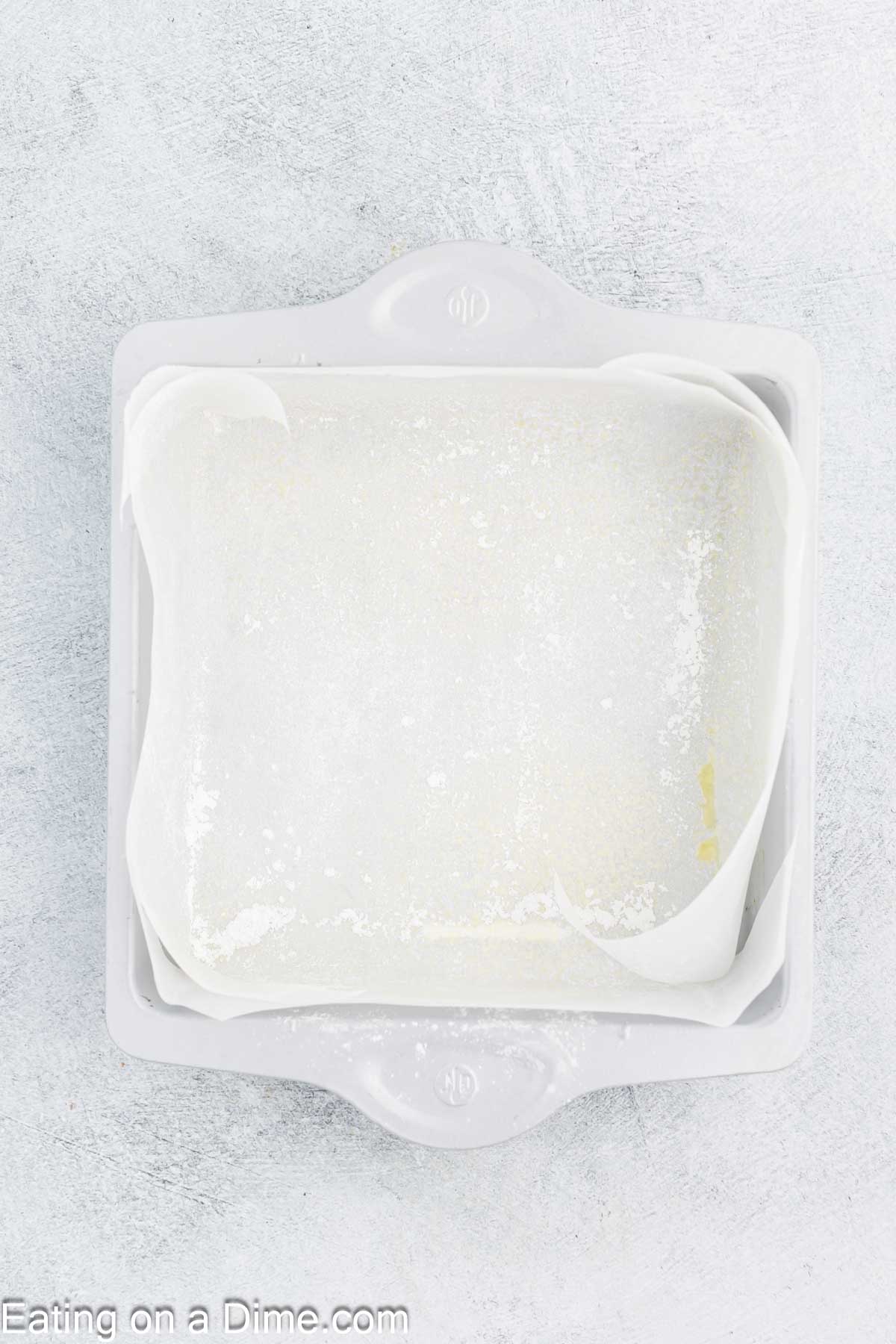 A square baking pan, ready for homemade marshmallows, is lined with white parchment paper and lightly greased, resting on a textured, light gray surface.