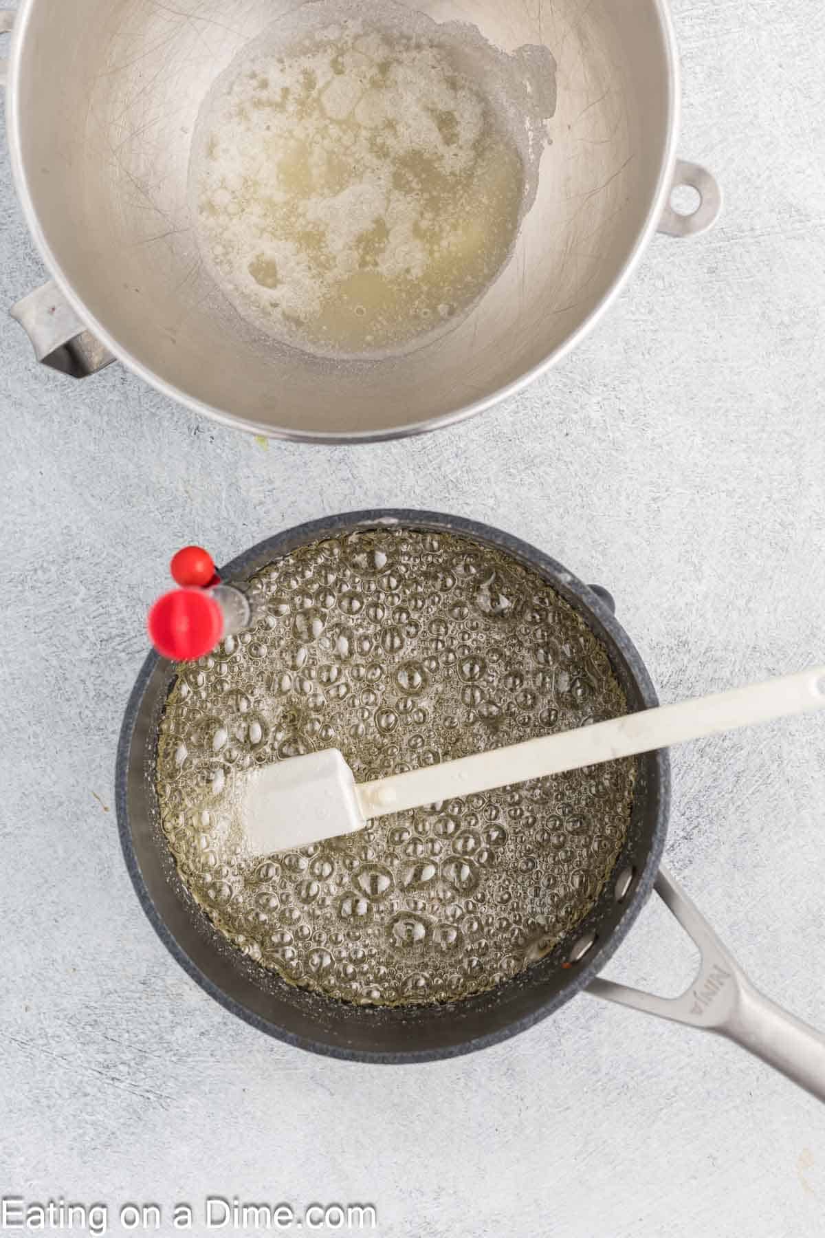 A top view of a saucepan filled with bubbling liquid for a marshmallow recipe and a white spatula resting inside. A separate metal bowl above contains a smaller amount of liquid, crucial for homemade confectionery. The background is a light gray surface.