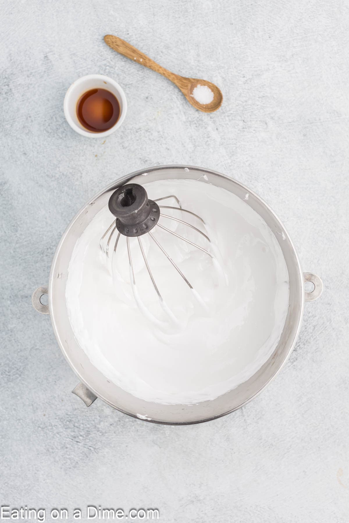 A mixing bowl with whipped egg whites and a whisk attachment awaits the magic touch needed for homemade marshmallows. Nearby, a small cup of vanilla extract and a wooden spoon with salt rest on a light gray surface. The text "Eating on a Dime.com" is at the bottom.