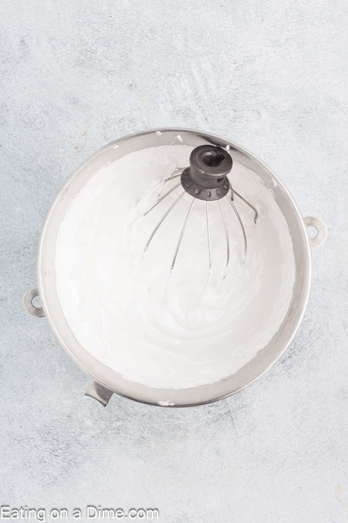 A metal mixing bowl filled with smooth, fluffy cream reminiscent of the beginnings of a marshmallow recipe, with a wire whisk attachment inside. The background is a light gray surface, perfect for crafting DIY marshmallows.