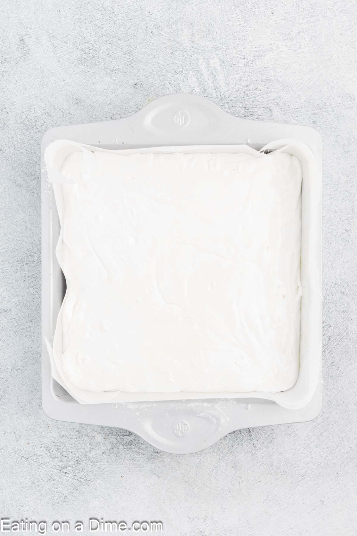 A square baking dish filled with a smooth layer of white frosting, perfect for layering your homemade marshmallows. The sides of the dish have small handles, and the background is a light gray textured surface.