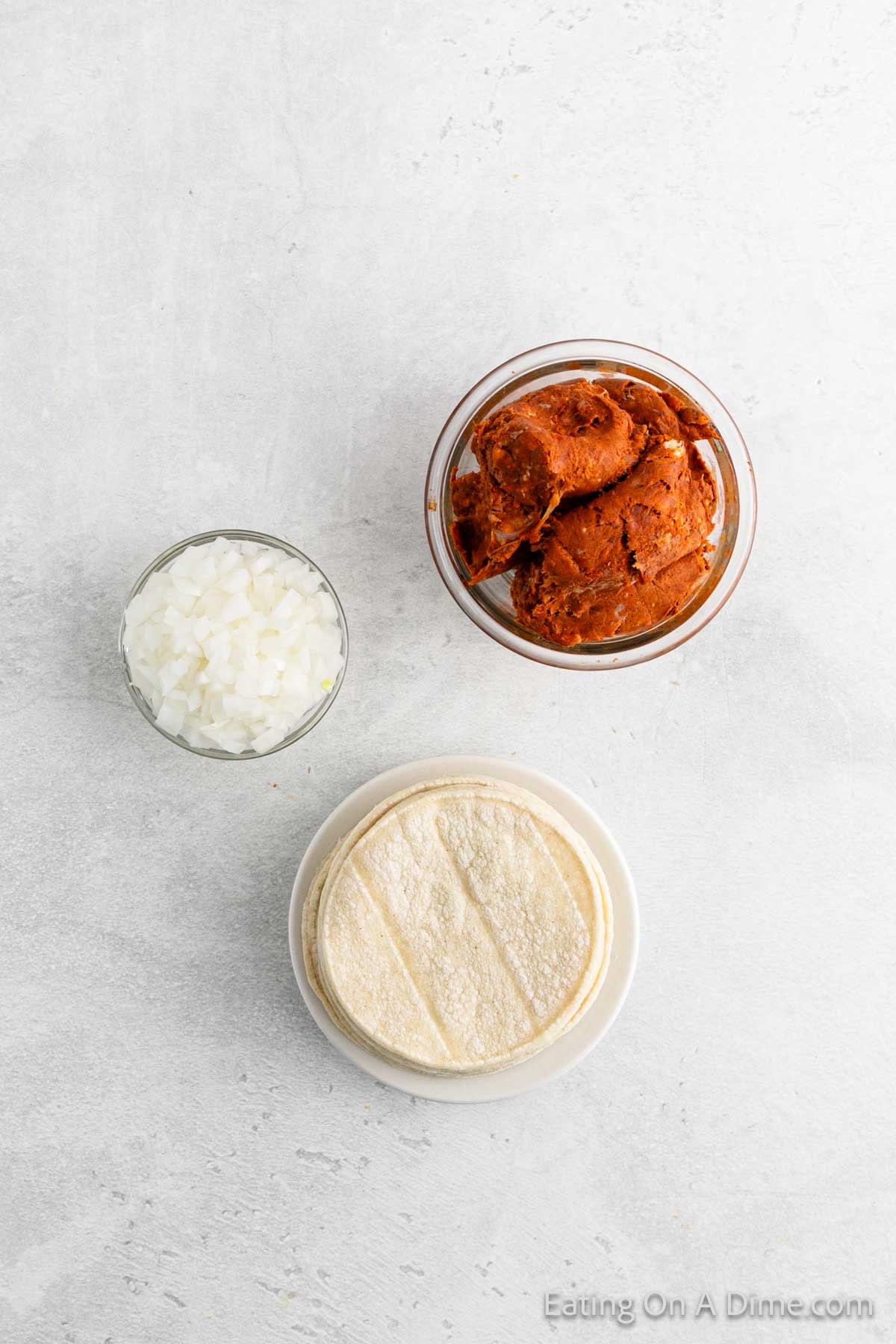 A bowl of refried beans, a small dish of chopped onions, and a stack of corn tortillas on a white surface await transformation into delicious street tacos.