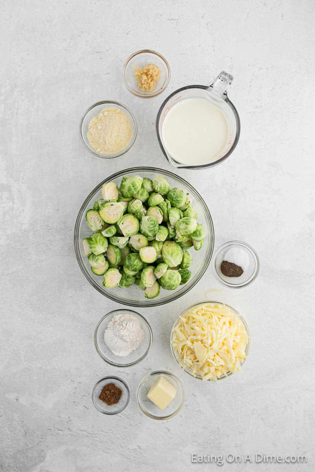 A top view of a cooking preparation scene for Brussels sprouts au gratin, featuring bowls filled with Brussels sprouts, milk, minced garlic, breadcrumbs, flour, shredded cheese, butter, ground black pepper, and nutmeg all beautifully arranged on a light gray surface.