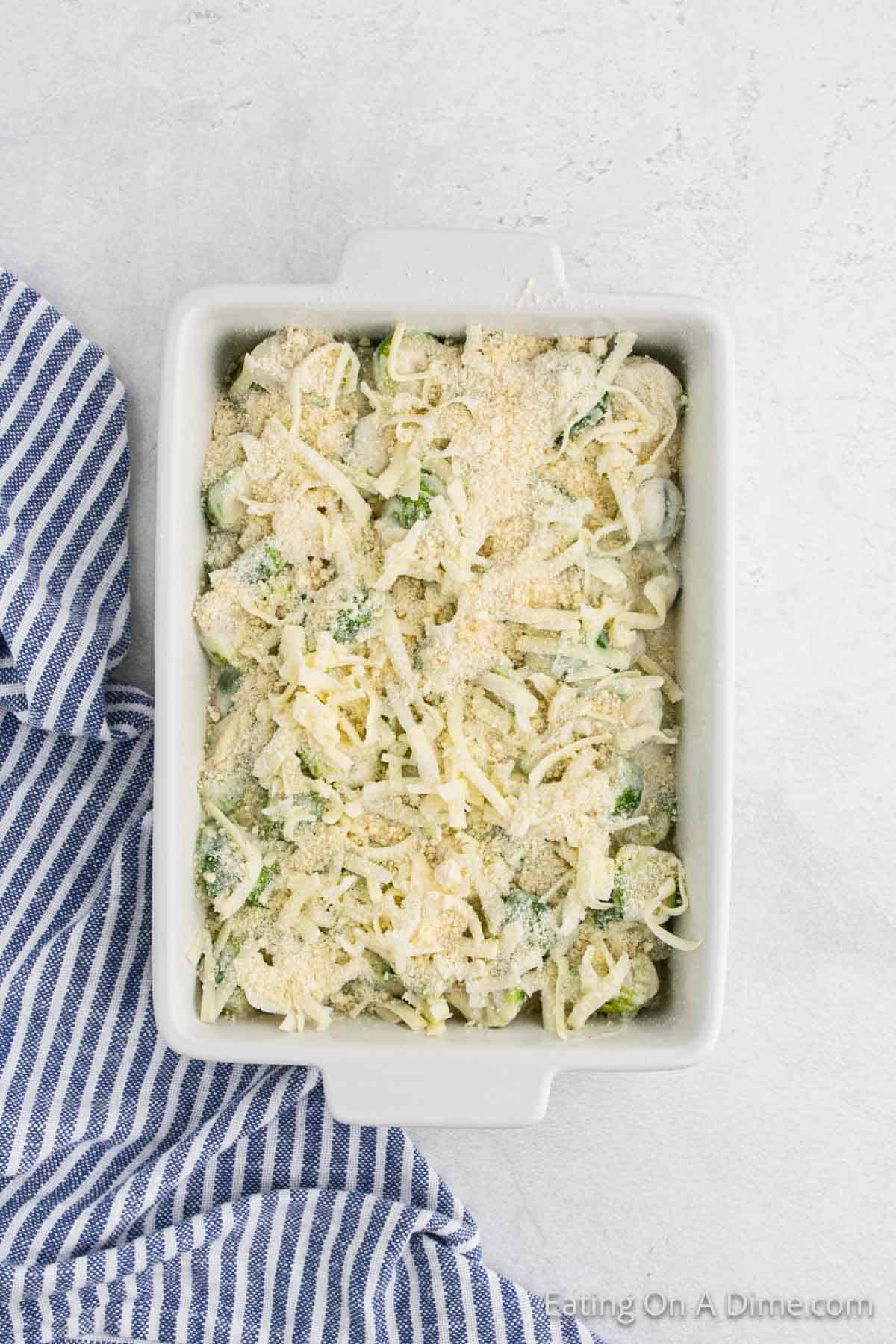 A rectangular white dish filled with a cheesy broccoli and cauliflower au gratin casserole, topped with melted cheese and breadcrumbs. A blue and white striped cloth is placed on the left side on a light gray marble surface, adding charm to this delectable recipe.