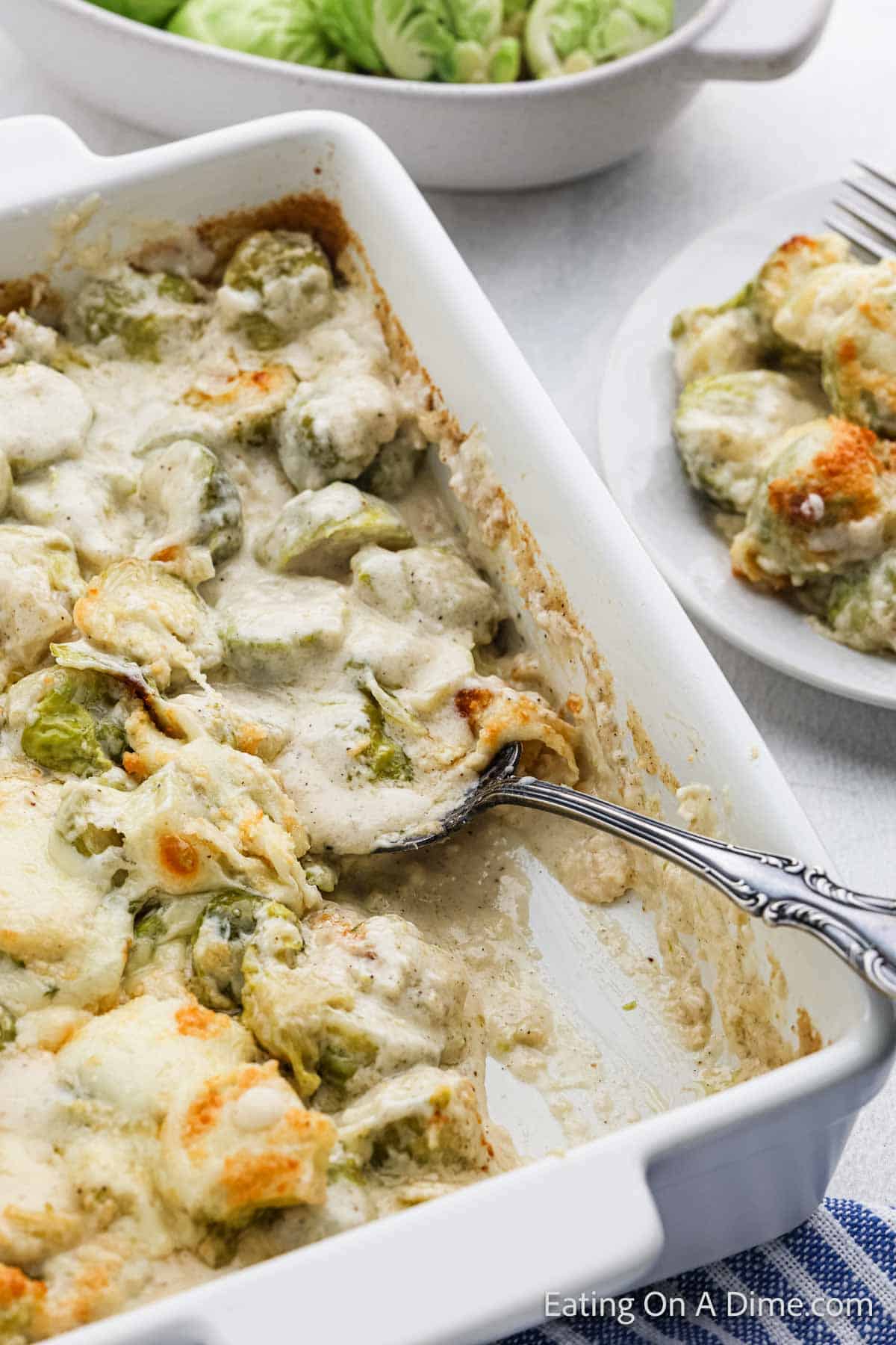 A close-up of an au gratin dish featuring brussels sprouts in a creamy cheese sauce, topped with melted cheese. A portion has been scooped out, revealing a silver spoon. A plate with a serving and a bowl of fresh brussels sprouts add to the recipe's appeal in the background.