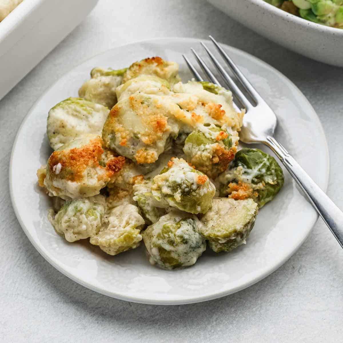 A plate of Brussels sprouts au gratin, topped with melted cheese and breadcrumbs, accompanied by a fork. This delightful recipe is served on a white plate against a light-colored background.