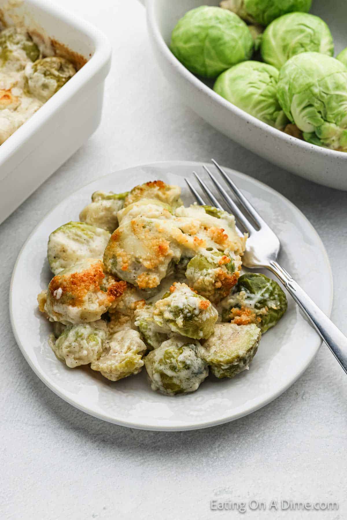 A plate of creamy Brussels sprouts casserole au gratin, topped with golden breadcrumbs, sits invitingly next to a fork. Nearby are a baking dish with more delicious casserole and a bowl of fresh Brussels sprouts, ready for your next culinary adventure. Perfect for any recipe collection!