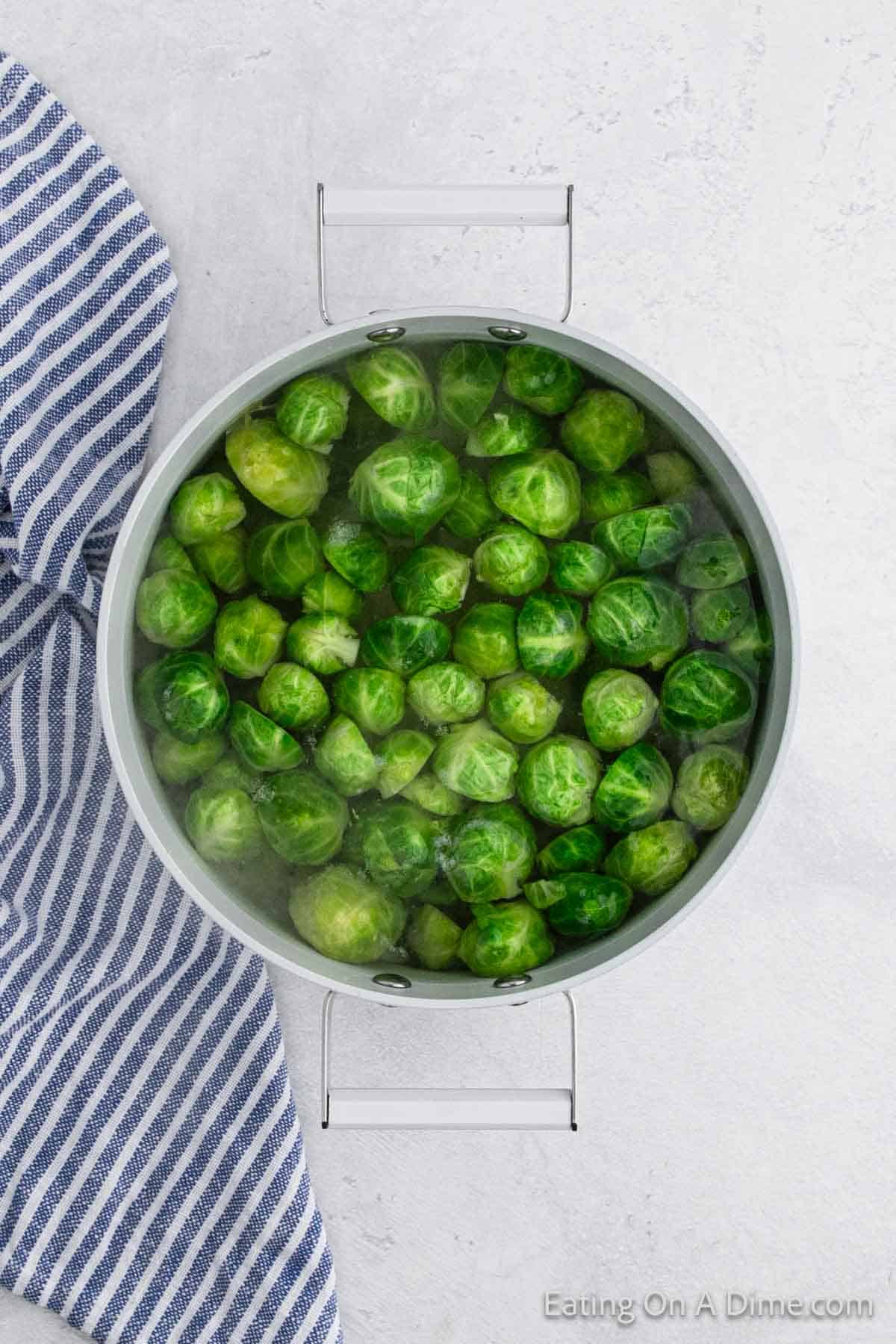 A pot filled with fresh green Brussels sprouts, ready to become a delicious Au Gratin dish, sits on a light-colored surface next to a blue and white striped kitchen towel. Steam gracefully rises from the pot.