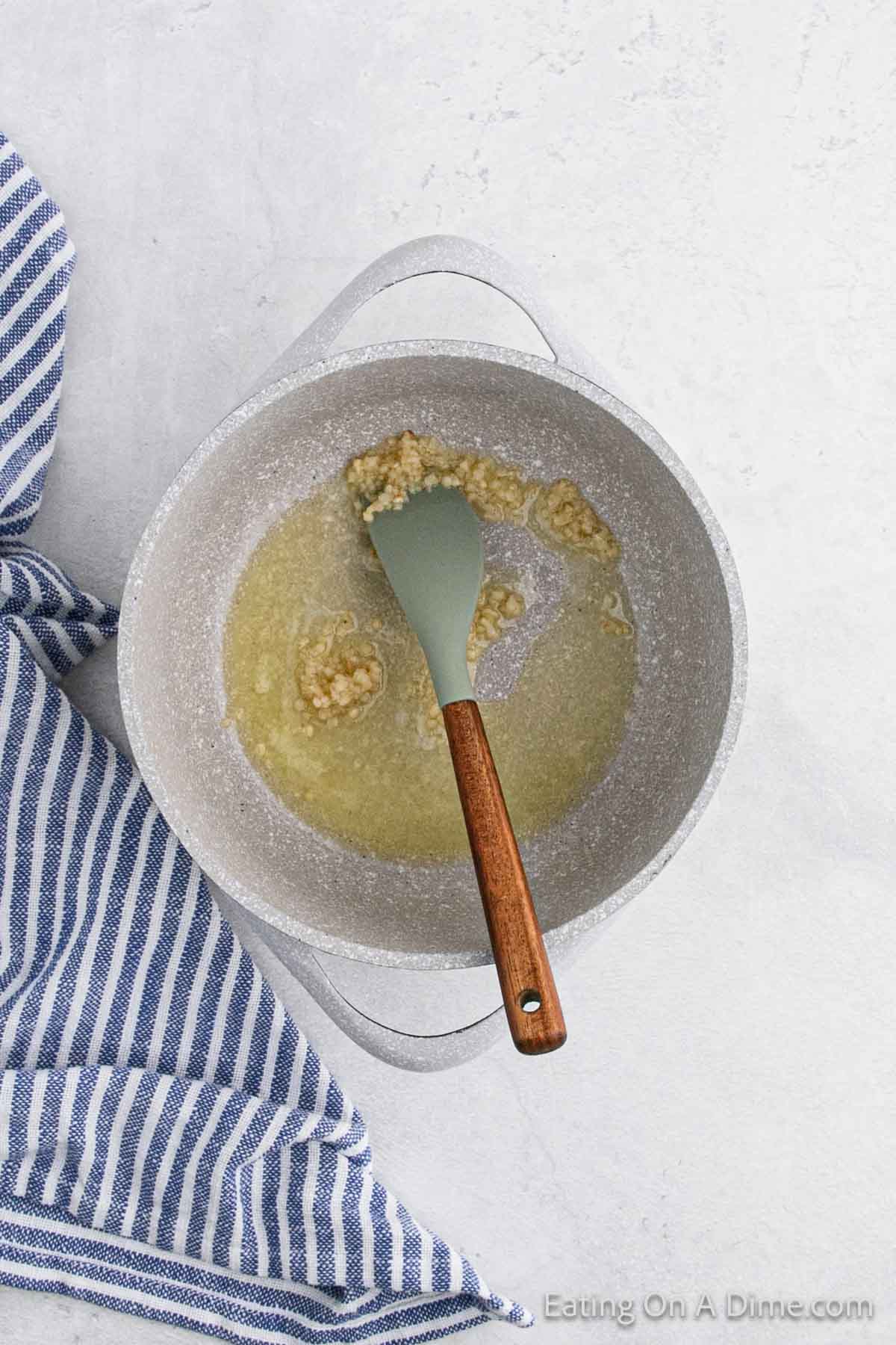 A pot with a wooden-handled spatula stirs minced garlic sautéing in butter, the perfect start for an Au Gratin recipe. A blue and white striped cloth lies beside the pot on a light surface, ready for the addition of tender brussel sprouts to elevate this delightful dish.