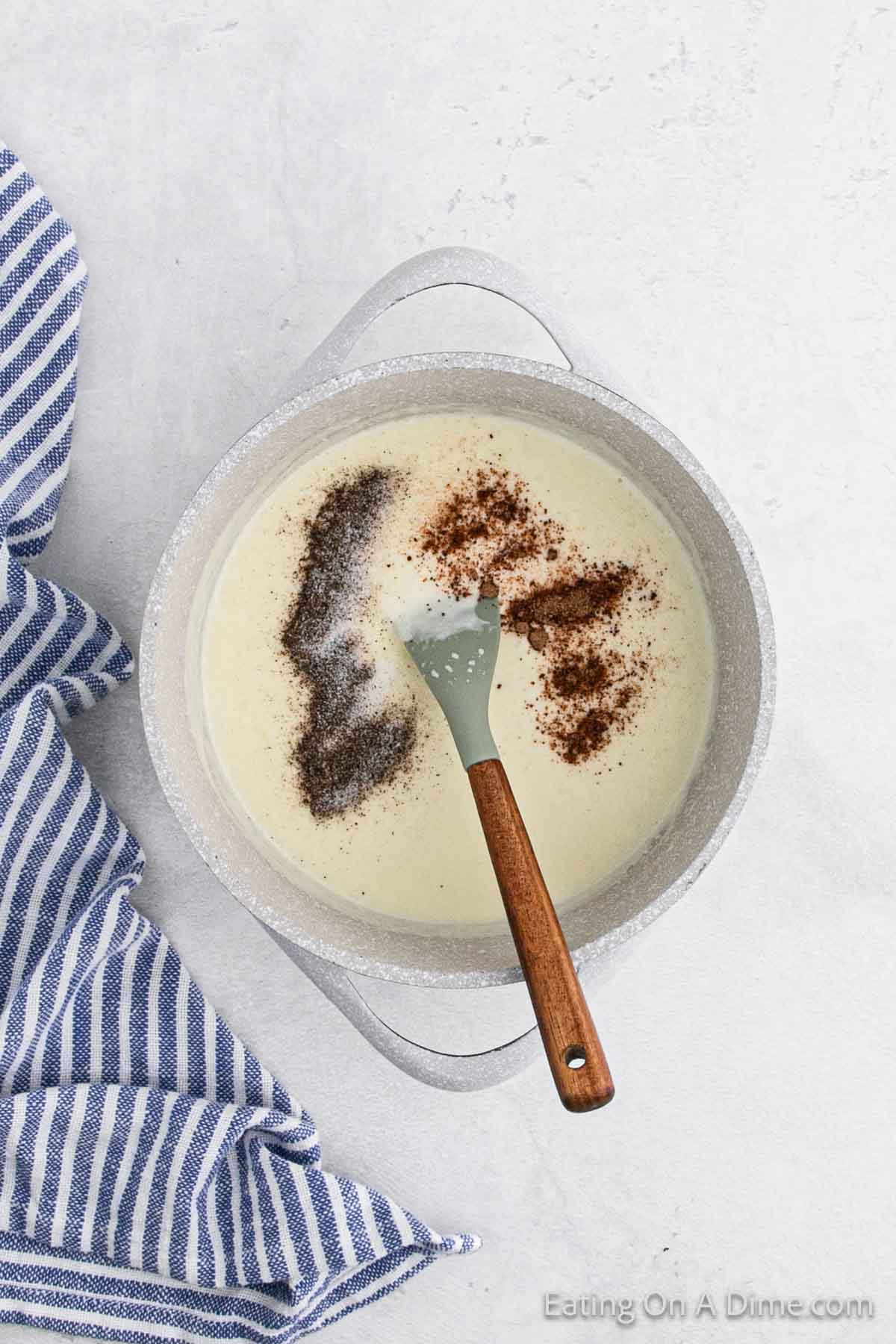 A pot of creamy sauce is being stirred with a wooden-handled spoon, perfect as a base for a Brussel Sprouts au gratin. The sauce, sprinkled with black pepper and other seasonings, sits beside a blue and white striped towel on a light gray surface.