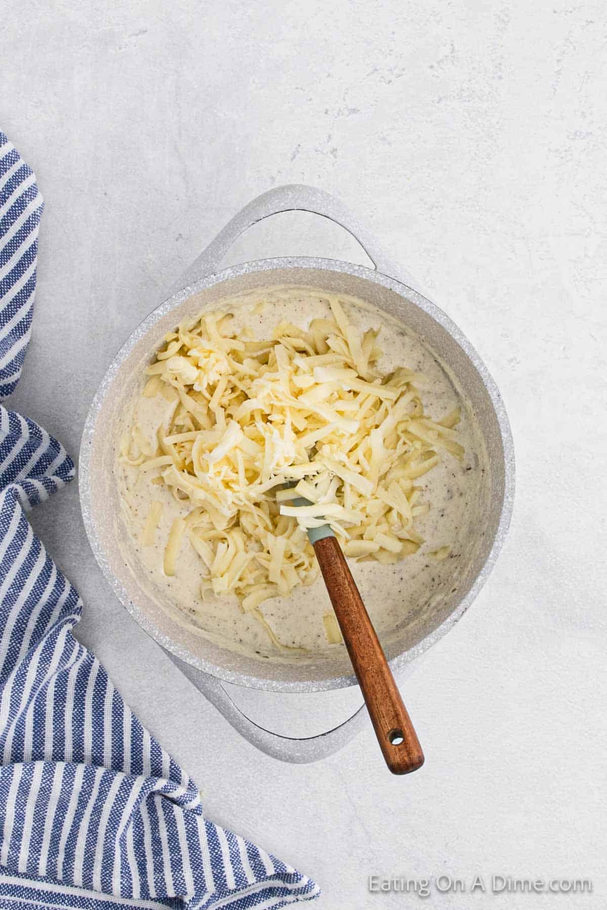 A pot filled with creamy sauce and shredded cheese, perfect for a Brussels sprouts au gratin recipe, with a wooden spoon resting inside. A blue and white striped cloth is on the left, placed on a light gray surface.